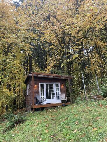 Una acogedora cabaña de madera se asienta en una ladera cubierta de hierba, rodeada de vibrantes árboles otoñales. Dos sillas descansan en el porche, invitando a relajarse en el abrazo de la naturaleza.