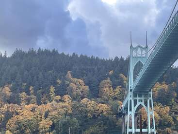 Un majestuoso puente colgante verde se extiende a través de la imagen, con exuberantes colinas boscosas que muestran los colores del otoño debajo, y un dramático cielo nublado dominando el fondo.