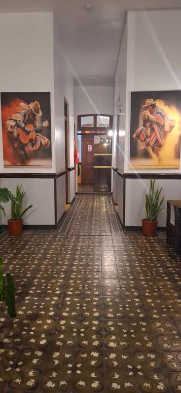 Um corredor com azulejos marrons estampados, vasos de plantas e duas pinturas vibrantes de músicos com roupas coloridas. Portas de madeira são visíveis no final.