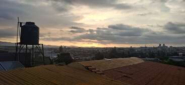 Essa imagem mostra uma vista da cidade ao pôr do sol, com nuvens incríveis no céu. Tem um tanque de água em cima de um telhado, com vista para vários prédios e montanhas distantes.