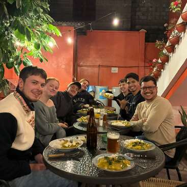 Sete amigos se juntaram em torno de uma mesa num ambiente aconchegante ao ar livre, compartilhando uma refeição alegre. A iluminação aconchegante, as plantas e os pratos de comida criam uma atmosfera acolhedora.