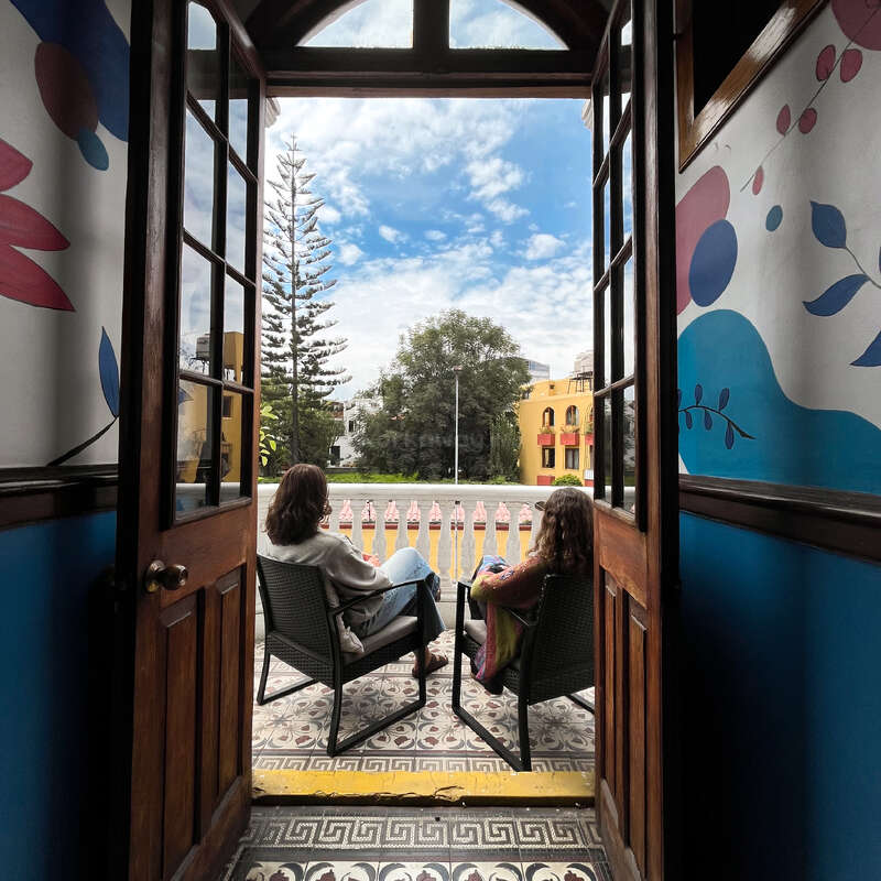Dos personas se relajan en un pequeño balcón, disfrutando de un día soleado. Las paredes interiores de colores vivos, las puertas de madera abiertas, las baldosas estampadas y unas vistas al exterior muy acogedoras completan la escena.