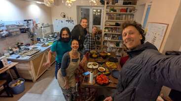 Six personnes sourient avec enthousiasme dans une cuisine confortable, autour d'une table remplie de divers plats. Des étagères contenant des épices et un tableau blanc décorent la pièce.