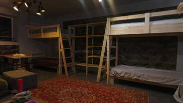 Cette chambre commune confortable comprend des lits superposés en bois, un tapis à motifs rouges, une table avec des bancs, des étagères, un ottoman gris et une atmosphère chaleureuse et accueillante.