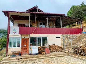 Maison rustique à deux étages avec un toit rouge, un tas de bois et un escalier extérieur. Entourée de verdure, elle dispose d'un balcon spacieux et d'une atmosphère simple et confortable.