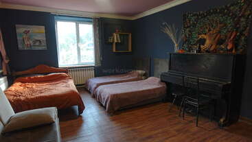 Cette chambre confortable dispose d'un lit, de deux matelas simples, d'une fenêtre, d'un tableau, de murs d'un bleu profond, d'un piano, d'une tapisserie décorative et d'un sol en bois.