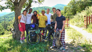 Un groupe de sept amis et un chien posent ensemble en plein air près d'un arbre et d'un vélo, avec des collines verdoyantes et un paysage de montagne ensoleillé en arrière-plan.