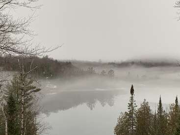 L'image représente une scène de lac sereine avec une atmosphère brumeuse, entourée d'arbres et d'un ciel gris, évoquant un sentiment de calme et de tranquillité.