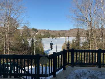 L'image représente une scène hivernale sereine depuis une terrasse, avec un lac gelé, des arbres et des mangeoires pour oiseaux, le tout dans un ciel bleu limpide.