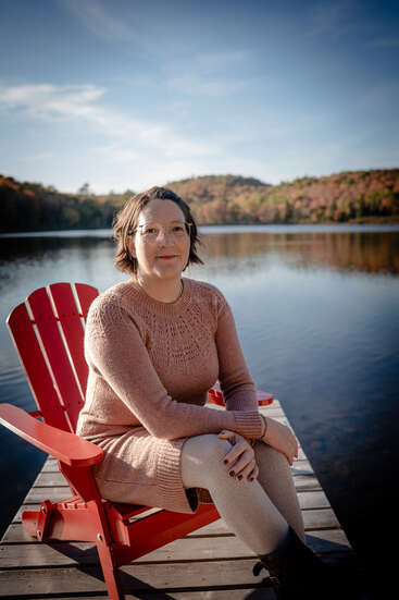 A woman sits on a red Adirondack chair on a lakeside dock, wearing a pink sweater dress and glasses, with autumn trees and calm water behind her.
