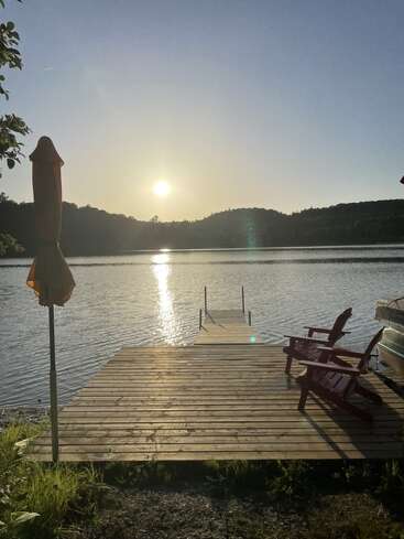 A peaceful lakeside scene with a wooden dock, two empty chairs, and umbrellas at sunrise or sunset, calm water, gentle light, and tranquil surroundings.