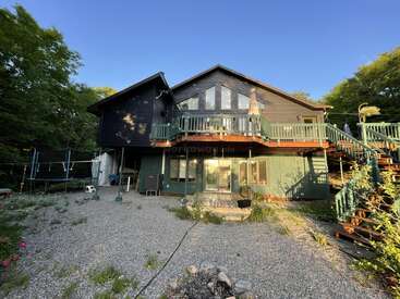 A rustic two-story house with dark wood siding, large windows, and wrap-around deck, nestled among lush green trees. Gravel yard, trampoline, and outdoor seating visible.