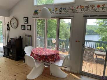 A bright dining area with modern white chairs, a colorful tablecloth, educational decorations, large windows, and a beautiful view of a lakeside deck and greenery outside.