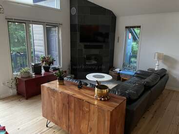 This cozy living room features a black sectional sofa, wooden furniture, large windows, indoor plants, a fireplace with TV above, and a serene, modern decor style.