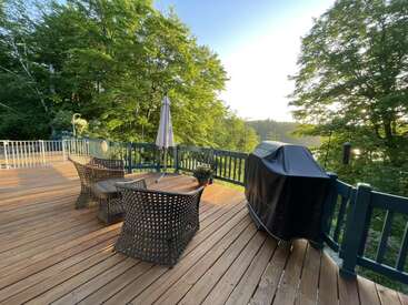 This image shows a sunny wooden deck with wicker chairs, a table, umbrella, and barbecue grill. Surrounded by trees, it's a peaceful outdoor setting.