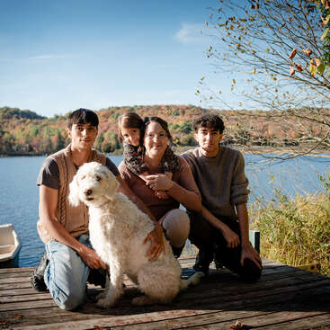 Uma família de quatro pessoas com um cachorro branco e fofinho posa em um deck de madeira à beira de um lago, cercada por árvores outonais sob um céu azul claro.