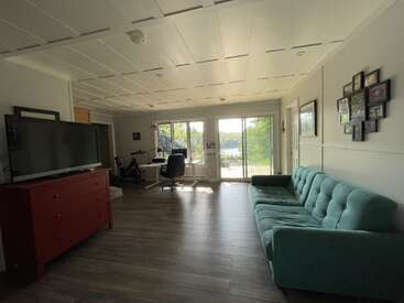 This living room features a teal sofa, red TV stand, flat-screen TV, office desk, large windows, sliding glass door, wooden floor, and family photos on the wall.