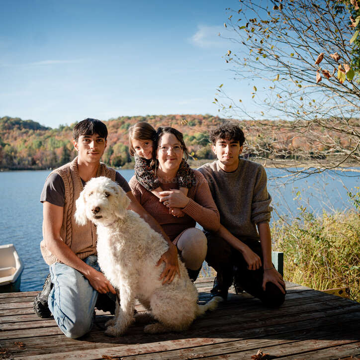A family of four with a fluffy white dog poses on a wooden deck by a lake, surrounded by autumn trees under a clear blue sky.
