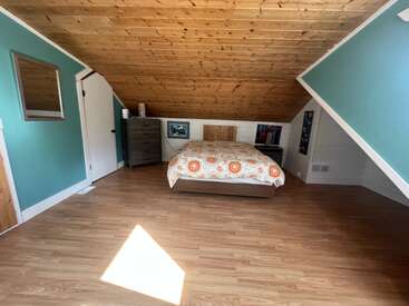 Dieses gemütliche Schlafzimmer im Dachgeschoss besticht durch schräge Holzdecken, blaugrüne Wände, ein Bett mit bunter Bettdecke, eine Kommode, Wandbilder und viel Tageslicht.