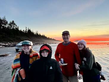 Vier Menschen lächeln und posieren gemeinsam am Strand bei Sonnenuntergang. Einige tragen Hüte und warme Jacken. Im Hintergrund sind ein Auto und Bäume zu sehen.