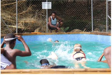 Plusieurs enfants nagent avec enthousiasme dans une petite piscine extérieure tandis qu'un adulte, accroupi au bord, les observe attentivement et les encourage. En arrière-plan, une clôture et une lumière du jour éclatante.