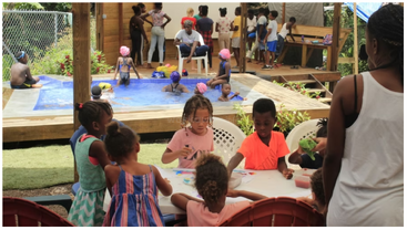 Des enfants sont réunis lors d'une animation en plein air très animée. Certains nagent dans la piscine tandis que d'autres sont assis à des tables où ils dessinent et peignent, sous la surveillance de plusieurs adultes attentifs.
