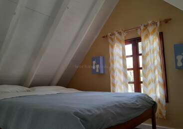 Cette chambre mansardée et chaleureuse est dotée d'un plafond blanc en pente, d'un grand lit recouvert d'une couverture bleue, de rideaux à motifs chevrons jaunes, de fenêtres en bois et d'une décoration murale ludique représentant des empreintes de pas. La pièce est baignée de lumière naturelle.