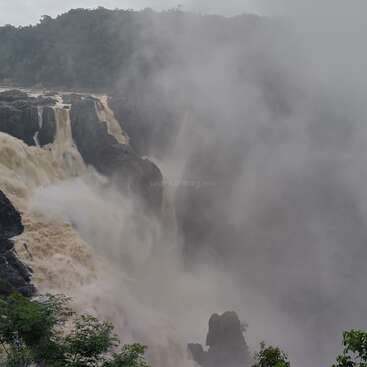 A powerful waterfall cascades over rugged cliffs, surrounded by dense mist and lush green foliage. The forceful water creates dramatic, cloudy spray against a forested backdrop.