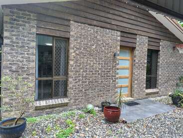 This image shows a brick house with two windows and a wooden door. There are potted plants, stones, and a basketball hoop on the right.