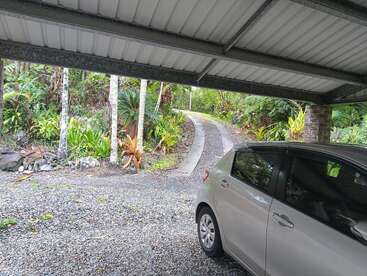 A silver car is parked under a metal carport. Beyond is a gravel driveway curving uphill, surrounded by lush green tropical plants and trees. Peaceful scene.