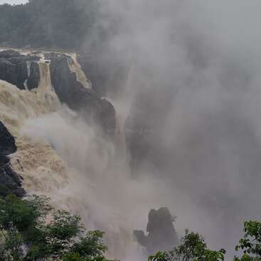 La brume s'élève d'une puissante cascade qui plonge sur des falaises sombres, entourées d'une végétation luxuriante. Le ciel est couvert, ce qui ajoute une atmosphère dramatique et mystérieuse.
