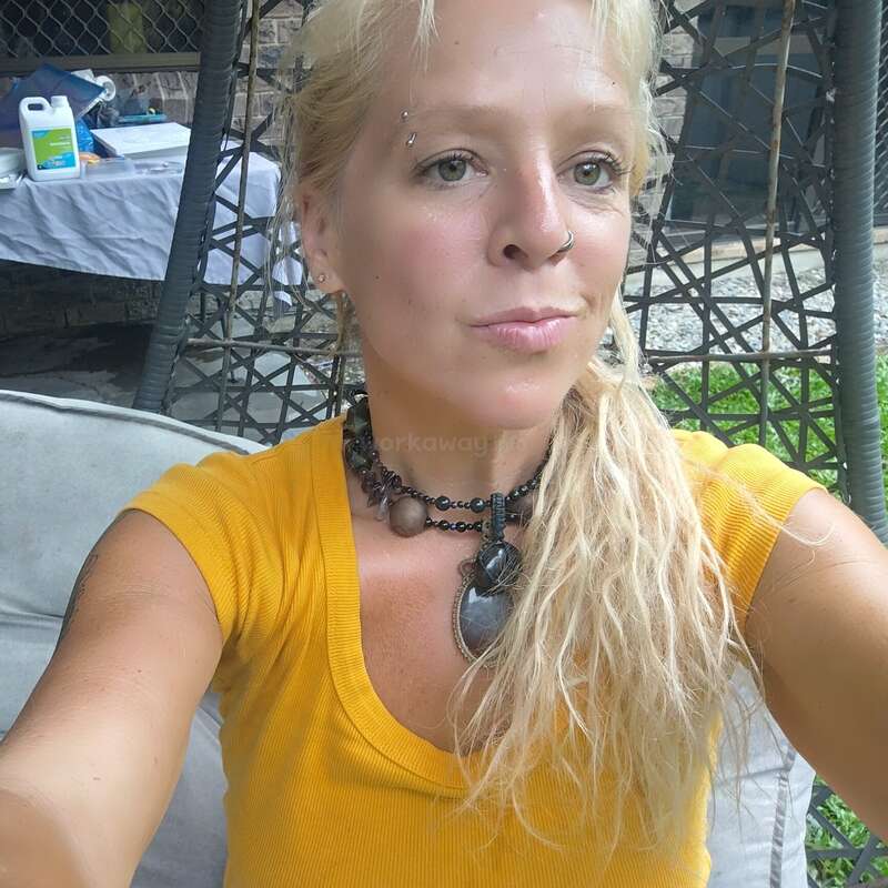A woman with blonde hair tied to the side sits outside on a chair, wearing a yellow shirt and layered necklaces, taking a relaxed selfie. Background shows greenery.