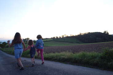 Trois enfants marchent main dans la main sur une route de campagne tranquille au coucher du soleil, entourés de champs et de collines ondulantes au loin, profitant ensemble d'une soirée paisible en plein air.