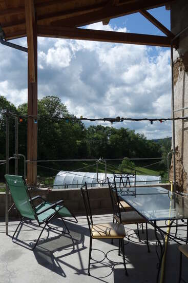 Une terrasse couverte avec une table et des chaises en verre, une chaise longue verte, des guirlandes lumineuses, donnant sur des arbres verdoyants, une serre et un ciel partiellement nuageux.