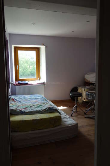 Une chambre simple et lumineuse, avec un matelas posé à même le sol, une literie colorée, une fenêtre, un parquet, des murs violet clair et une batterie dans un coin.