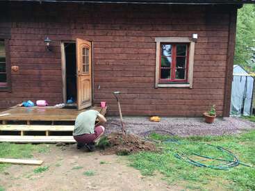 A person crouches in front of a wooden house, digging with a shovel. There are scattered objects, hoses, and a potted plant near the entrance steps.