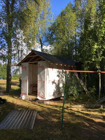 A small shed or cabin under construction stands among tall trees. Its exterior is wrapped in insulation material. Nearby, building materials and a swing set are visible.