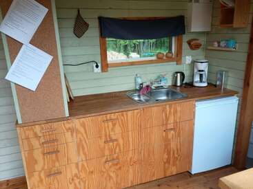 A cozy wooden kitchen features a countertop, drawers, sink, coffee maker, kettle, mini fridge, window with forest view, notes on corkboard, and minimal decor.
