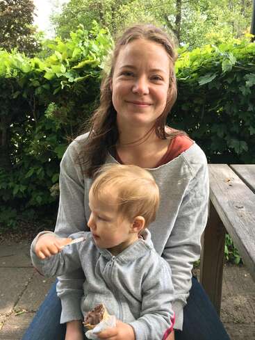 A smiling woman sits outside on a bench, holding a young child eating ice cream. Lush green bushes and trees surround them in the sunny background.