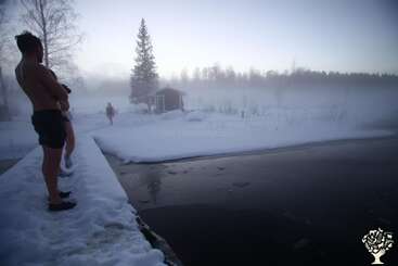 Three people in swimsuits stand by a snow-covered dock, preparing for a winter swim in a partially frozen lake, surrounded by mist, trees, and a small cabin.