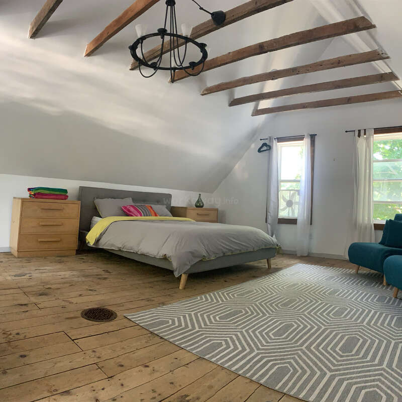 Esta acogedora habitación cuenta con vigas de madera a la vista, una cama moderna con almohadas de colores, cómodas de madera, una alfombra geométrica, grandes ventanas con cortinas transparentes y suelos de madera rústicos.