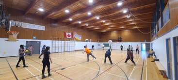 Eine Gruppe von Leuten spielt Völkerball in einer geräumigen Turnhalle mit Holzwänden und -decke. Fahnen und Basketballkörbe sind zu sehen und sorgen für eine lebhafte Atmosphäre.