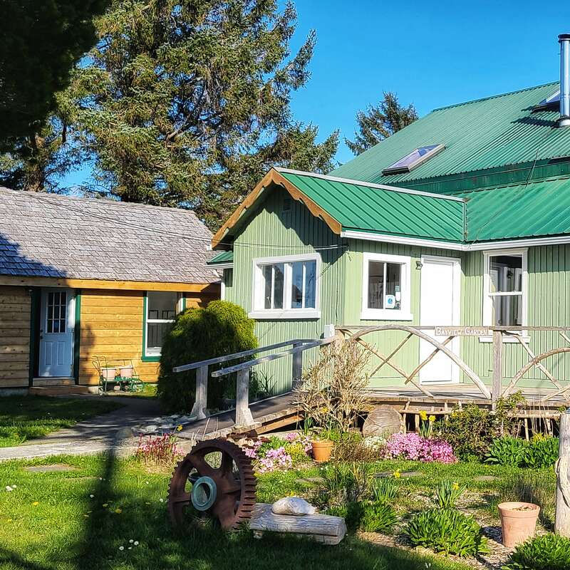 The image depicts a charming green house with a green roof, surrounded by a garden and a wooden bridge, set against a serene blue sky.