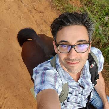 Un homme aux cheveux bruns courts et aux lunettes prend un selfie sur un chemin de terre, avec l'arrière d'un cheval brun visible derrière lui.