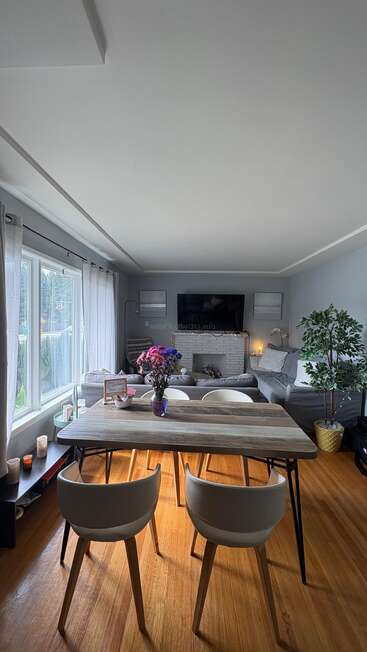 Uma sala de estar aconchegante e moderna, com janelas amplas, piso de madeira, uma mesa de jantar, duas cadeiras, flores frescas, uma TV, um sofá e plantas decorativas.