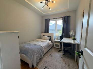 Este quarto aconchegante e minimalista tem uma cama de solteiro, uma cômoda branca, uma mesa de trabalho elegante com cadeira, iluminação moderna, uma janela com cortinas cinza e um pequeno vaso de plantas decorativo.