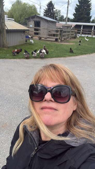 Uma mulher usando óculos escuros grandes e uma jaqueta preta tira uma selfie ao ar livre, com galinhas e edifícios da fazenda visíveis ao fundo, num dia nublado.