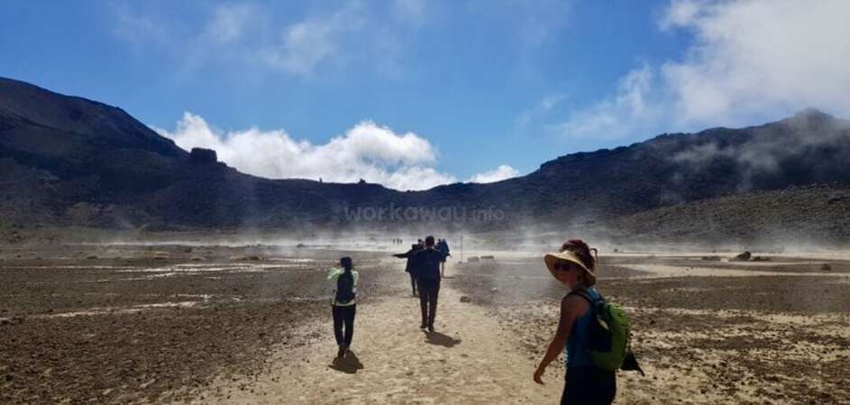 A group of three people walking along a sandy flat, a woman is turning back to smile at the camera. There are mountains and a blue sky in the distance.