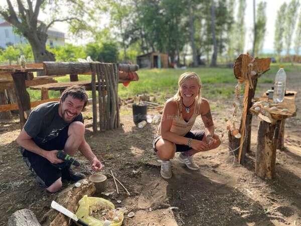 Two people crouching down in a sunny garden. The person on the left is holding a drill and appears to be using it to de-shell nuts. They are both dressed in summer clothes and have beaming smiles.