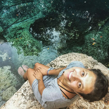 solo female traveller smiling at camera while sitting at the river with feet in water and fish swimming around them